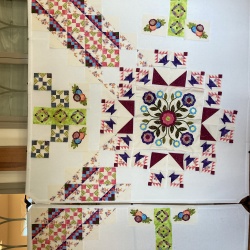This is what I’ve done so far, or at least the top half (the matching pieces would be on the floor).  I’m trying to decide on the colors for the cats cradle blocks—am thinking of the lighter blue for the 2 main triangles and pink for the outside point.  The smaller appliqué pieces aren’t done but the big one in the center is almost done.